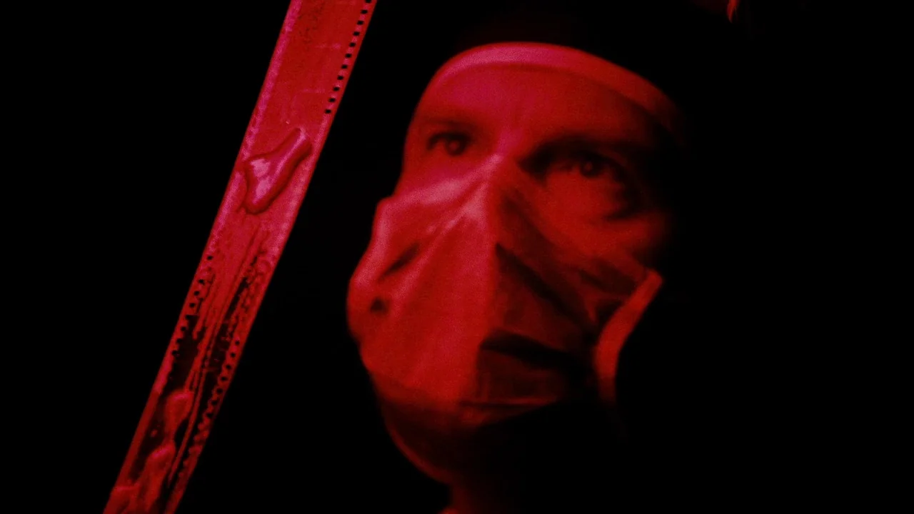 The ABCs of Death (2012) movie still: closeup — A person in a surgical mask stands in a darkened environment lit by red light. They hold a strip of…