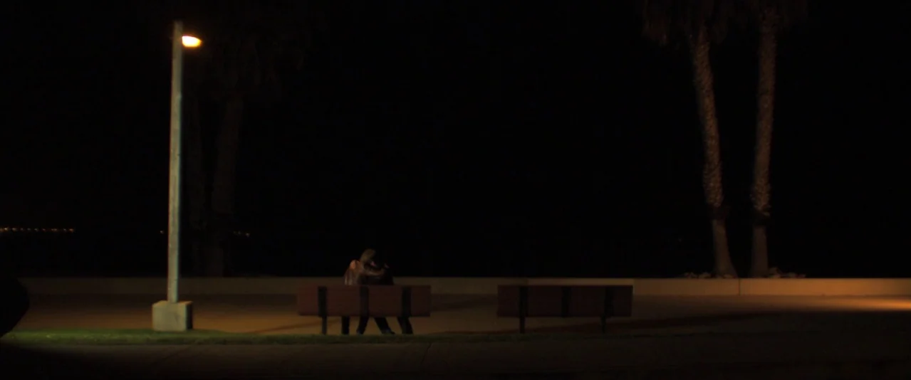 Bellflower (2011) movie still: wide — A dark outdoor area features two wooden benches placed along a paved walkway at night. Two people sit closely…