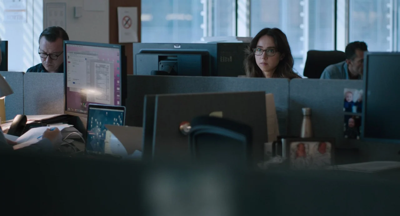 She Said (2022) movie still: medium — A modern open-plan workspace filled with grey fabric cubicles and computer equipment. A woman sits behind a…