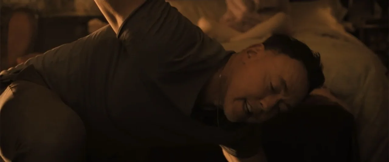 Abigail (2024) movie still: medium — A dimly lit bedroom features a neatly made bed and dark wooden furniture. A man hunches over near the side of…