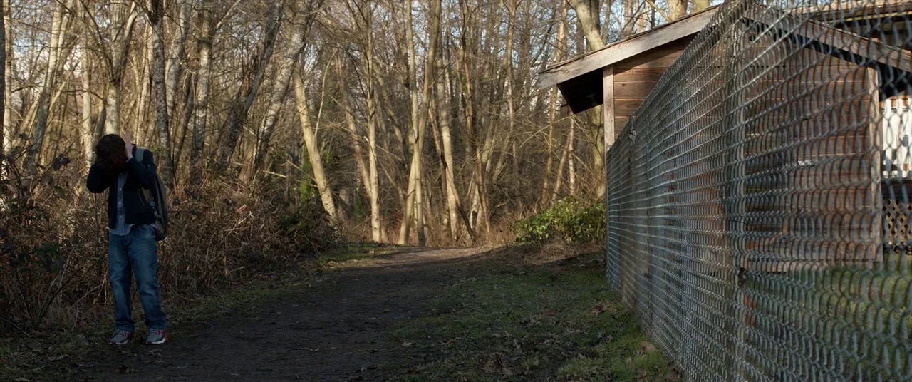The Predator (2018) movie still: wide — A dirt trail runs through a forest next to a tall chain-link fence. A young man stands alone on the path…