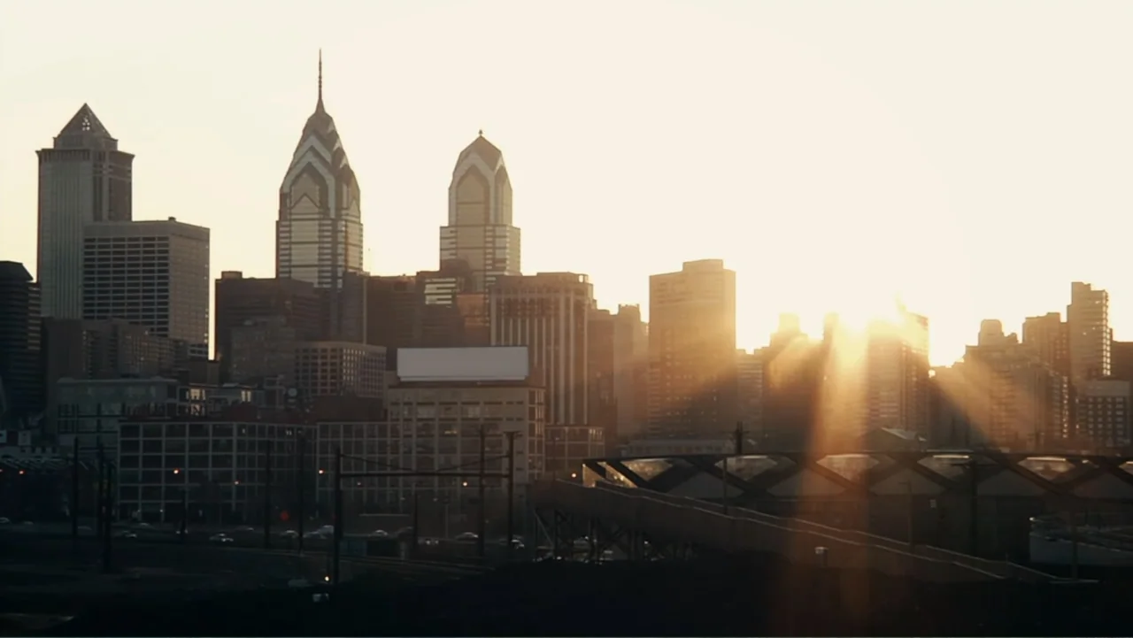 Toss It (2019) movie still: establishing — A dense metropolitan skyline sits under a clear sky during the golden hour. Sunbeams radiate across the…