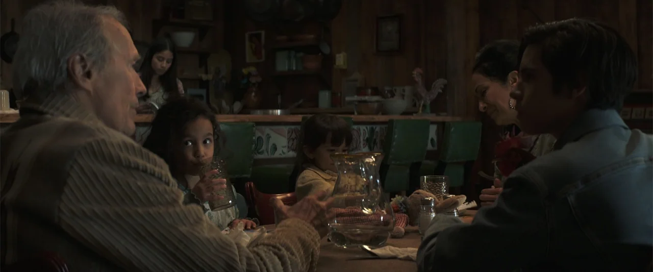 Cry Macho (2021) movie still: medium — A rustic dining room features wood-paneled walls and a table surrounded by family members. An elderly man in…