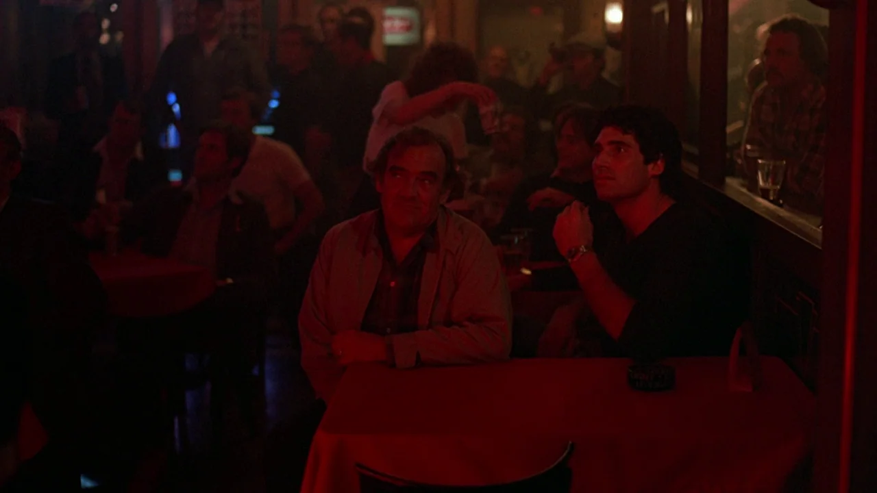 Flashdance (1983) movie still: medium — A dark tavern interior features red-clothed tables and dim ambient lighting conditions. Two men sit at a…