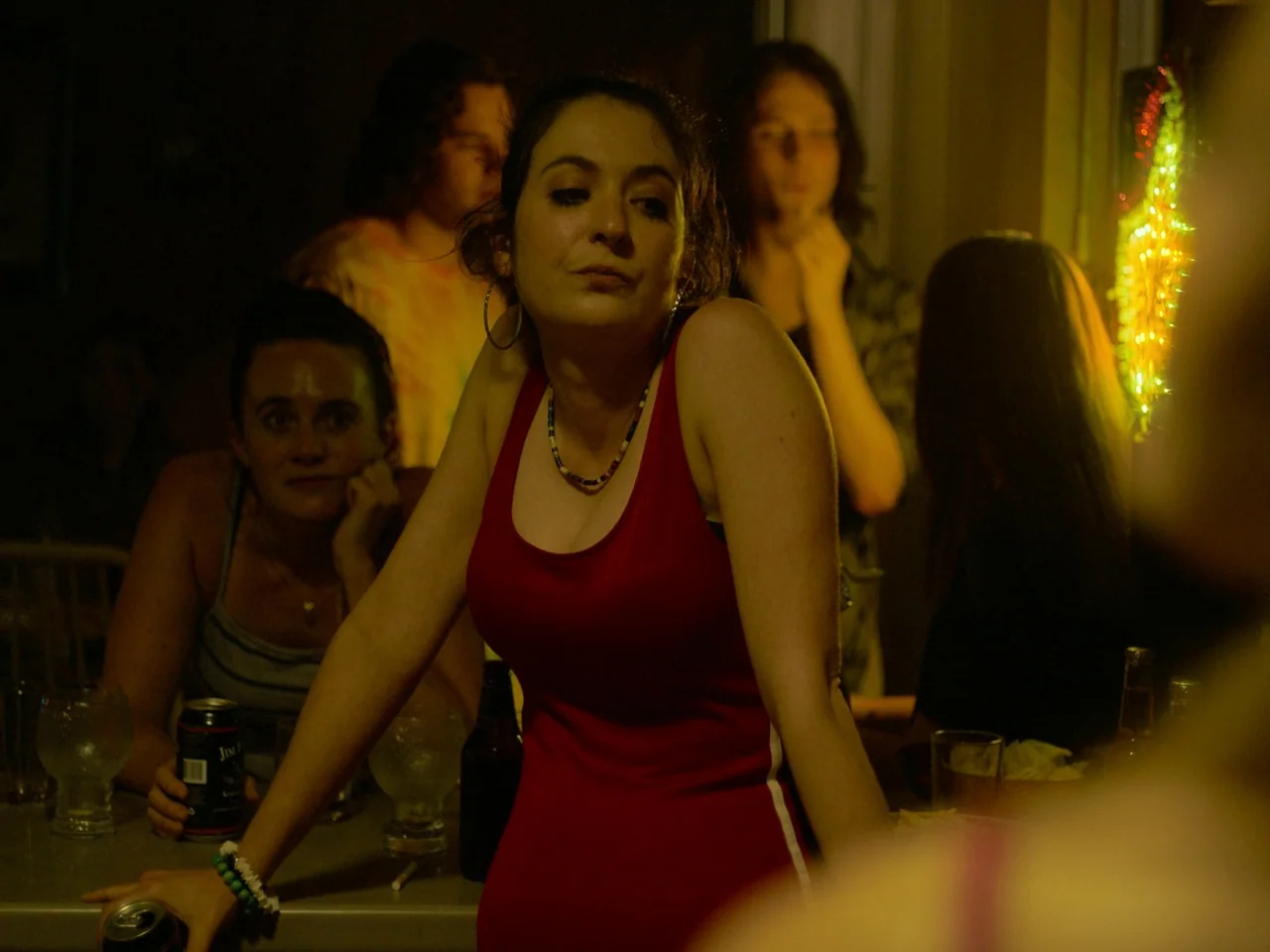 Of An Age (2022) movie still: medium — A dimly lit room populated by several young adults during a social gathering. A woman in a red top leans over…