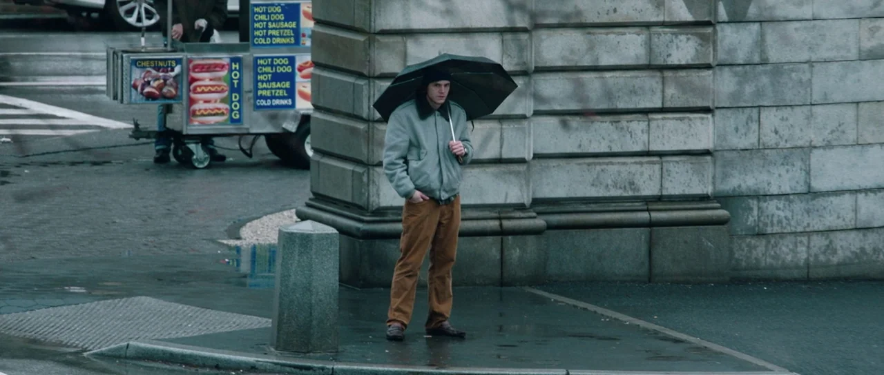 American Animals (2018) movie still: fullbody — A man stands on a grey concrete corner next to a stone building wall. A mobile food cart vendor is…