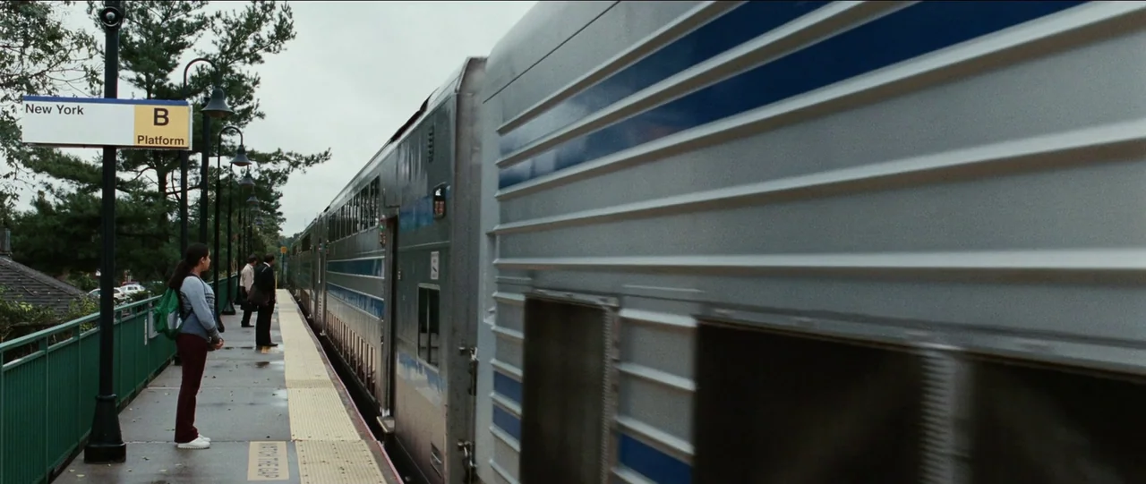 Bad Education (2019) movie still: wide — A train platform with a New York B Platform sign and metal safety railings. A young woman and two men stand…