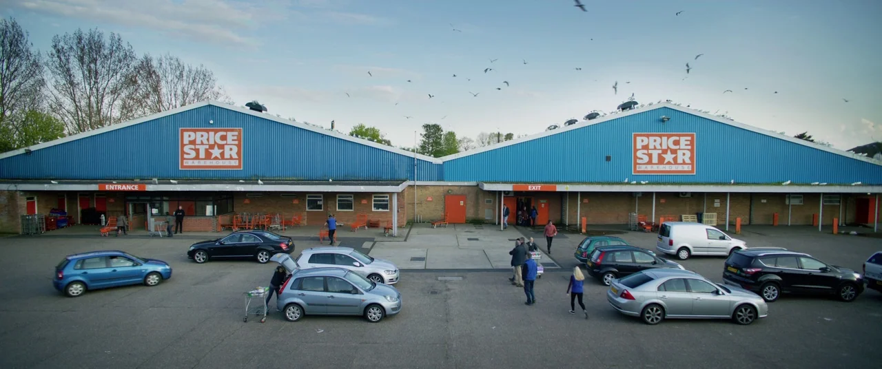 Yesterday (2019) movie still: establishing — A wide view of a warehouse store facade features dual blue triangular roof structures. Shoppers walk…