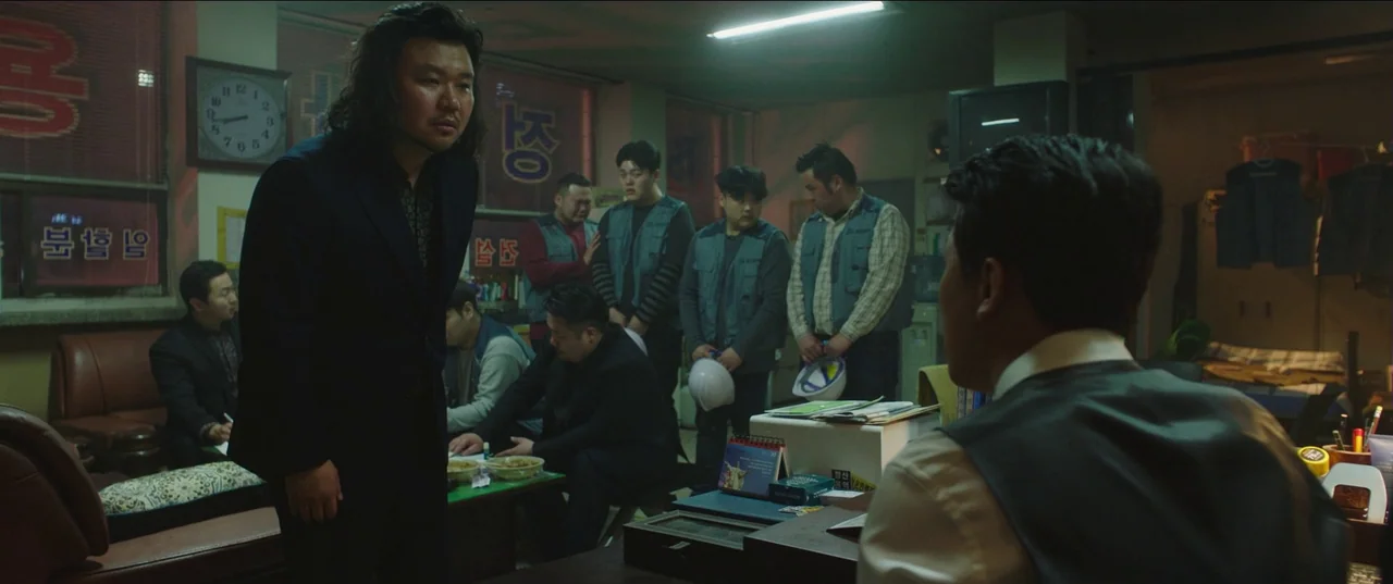 Psychokinesis (2018) movie still: medium — A cluttered room contains multiple men standing near a desk under fluorescent lighting. One man in a dark…
