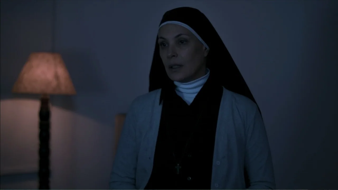 The Grotto (2022) movie still: medium — A dimly lit room with a single floor lamp glowing in the background. A woman dressed as a nun stands in the…