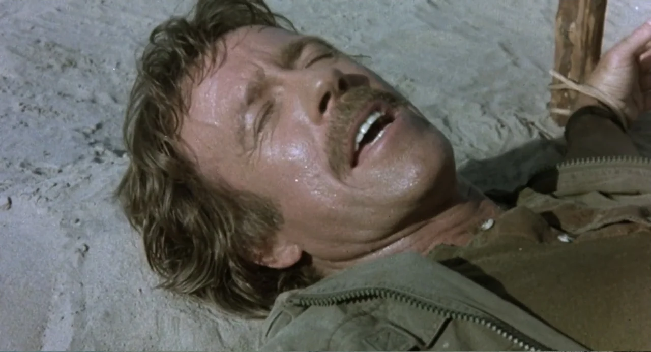 Firewalker (1986) movie still: closeup — A man lies on his back in the sand under bright direct sunlight. He wears a zipped jacket and has his…