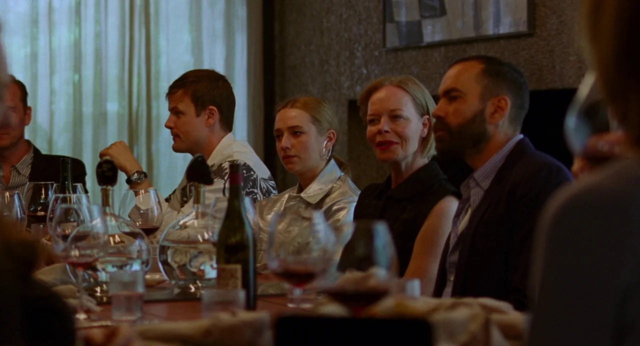Sick of Myself (2022) movie still: medium — A group of four adults sits around a dinner table with wine glasses. They appear to be engaged in…