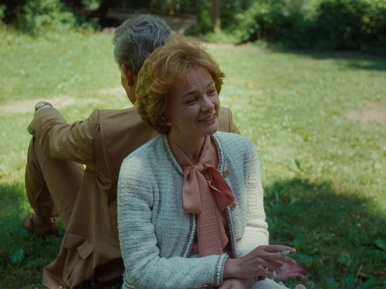 Maestro (2023) movie still: medium — A grassy lawn with blurred trees and shadows in the background during daytime. A woman with red hair smiles…