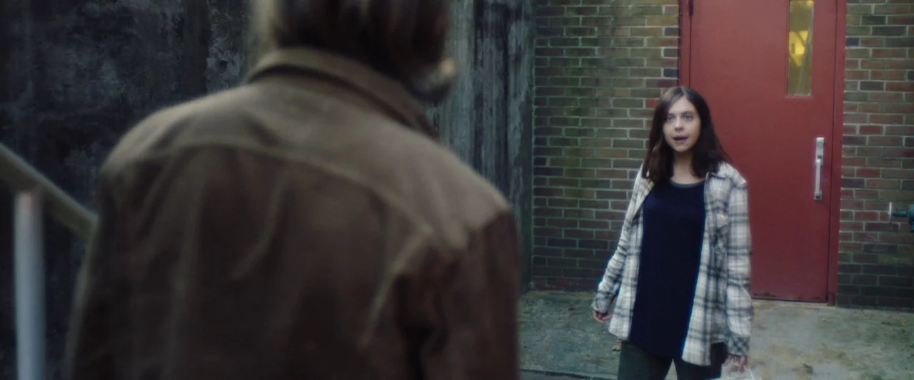 Wildling (2018) movie still: medium — An outdoor area features a brown brick wall and a prominent red industrial door. A young girl in a plaid shirt…