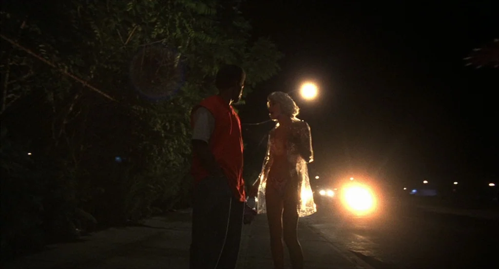 He Got Game (1998) movie still: fullbody — A dark, paved road at night with a dense tree line on the left. A man and a woman stand together on the…