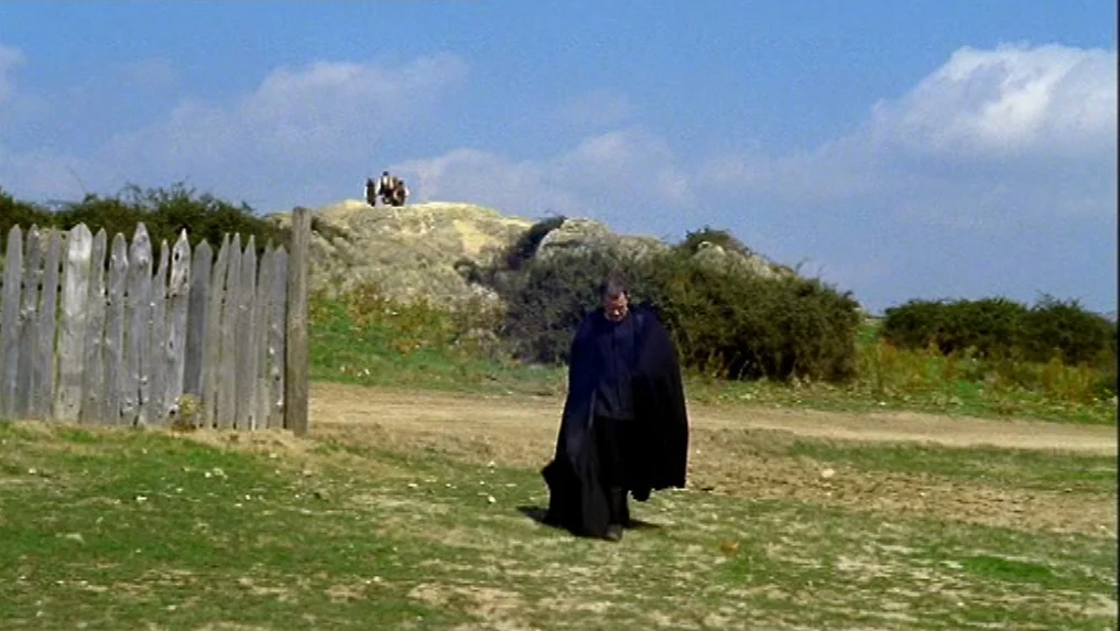 The Scarlet Letter (1973) movie still: wide — An open dirt field with a wooden stockade fence and a distant rocky mound. A man wearing a long black…