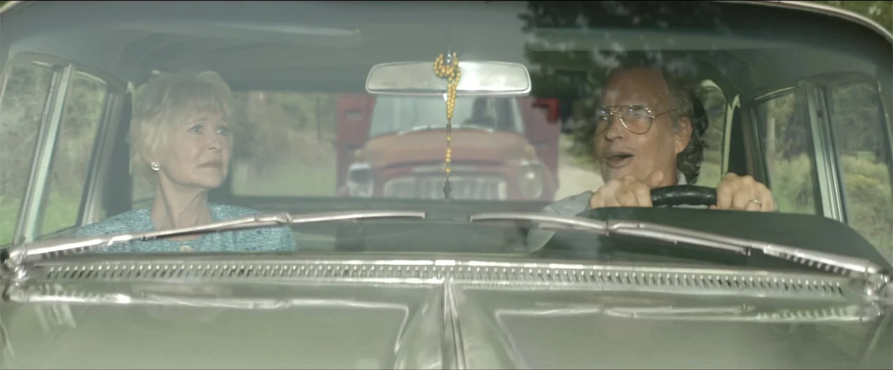 Jeepers Creepers: Reborn (2022) movie still: medium — An older couple travels inside a vintage car on a rural dirt road. A rusty truck follows…