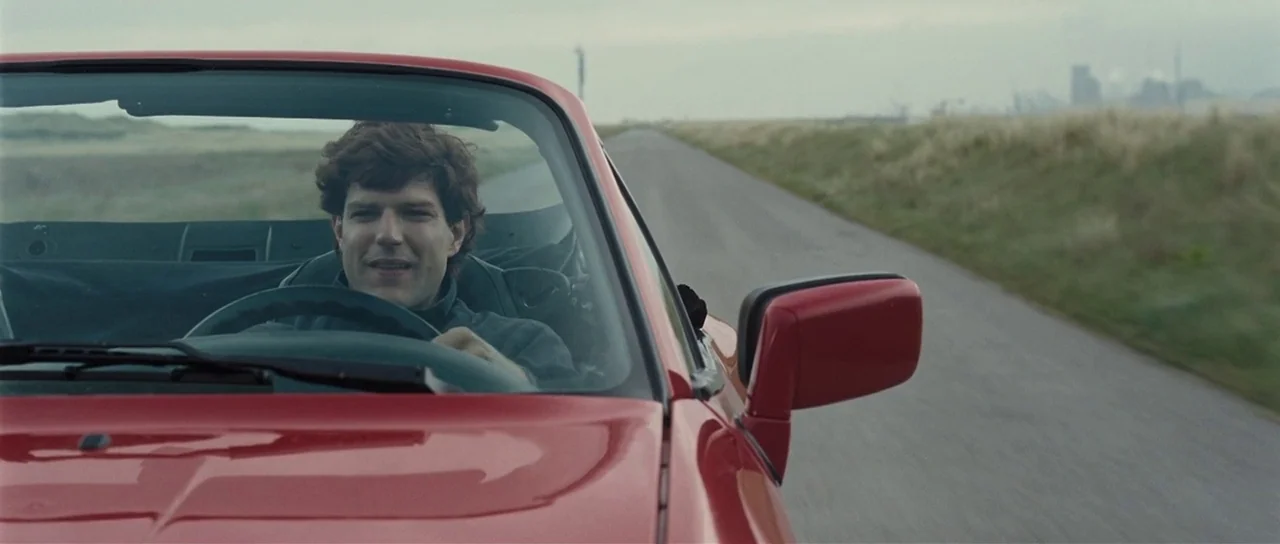 Our Day Will Come (2010) movie still: medium — A man drives a red convertible on an asphalt road through a grassy field. An industrial landscape is…
