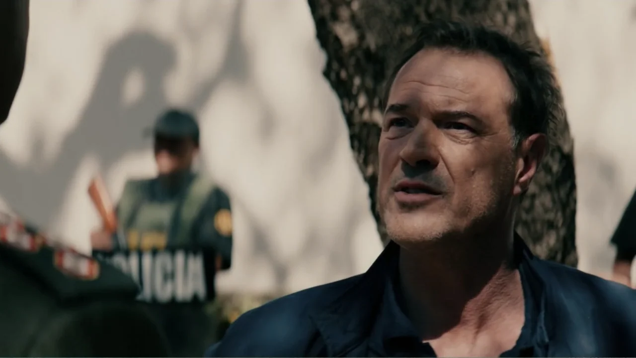 Bel Canto (2018) movie still: medium — An outdoor area features a large tree and a background police officer. A man with short dark hair speaks…