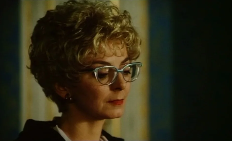 Simon, The Magician (1999) movie still: closeup — A woman stands indoors against a wall with vertically striped light and dark wallpaper. She looks…