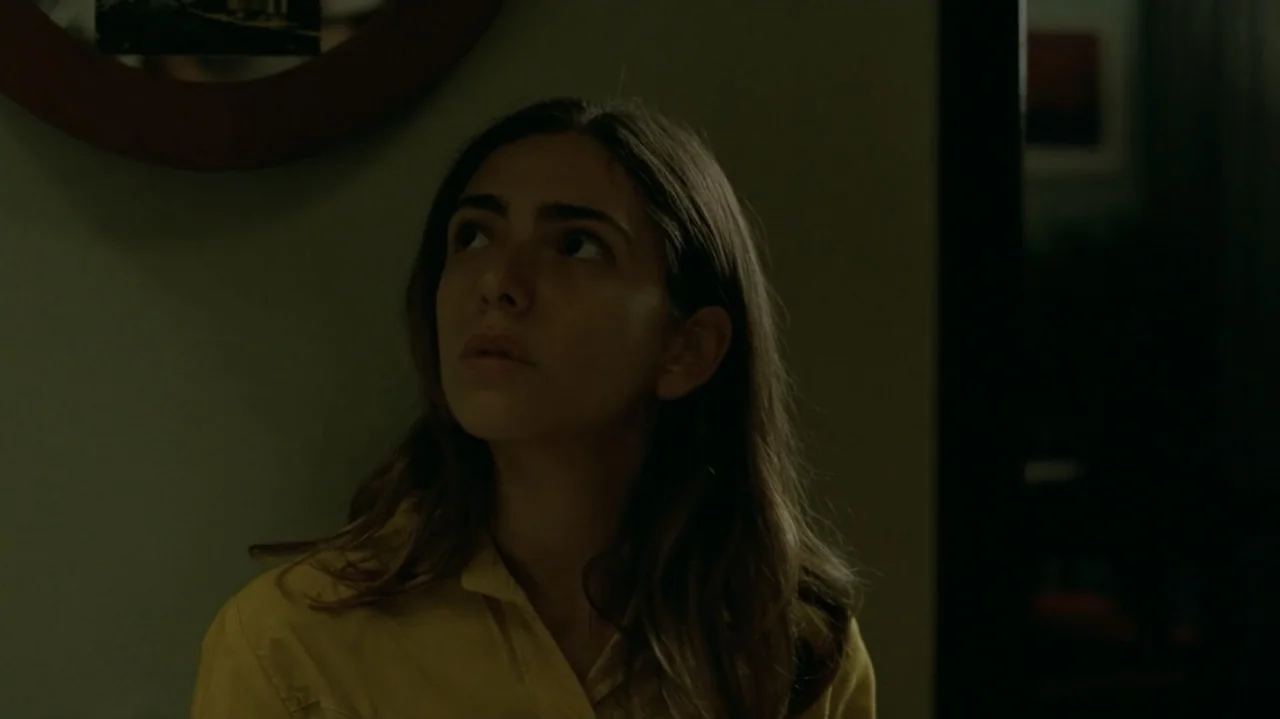 I'm Still Here (2024) movie still: medium — A dimly lit indoor space with plain wall surfaces and minimal background decor. A woman with long brown…