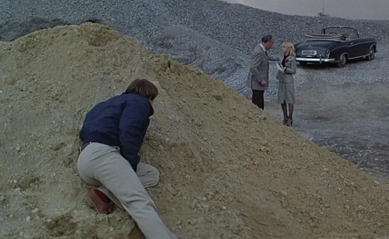 Maîtresse (1975) movie still: wide — A gravel and sand pit features large mounds of stone aggregate. A man hides behind a sand hill while an older…