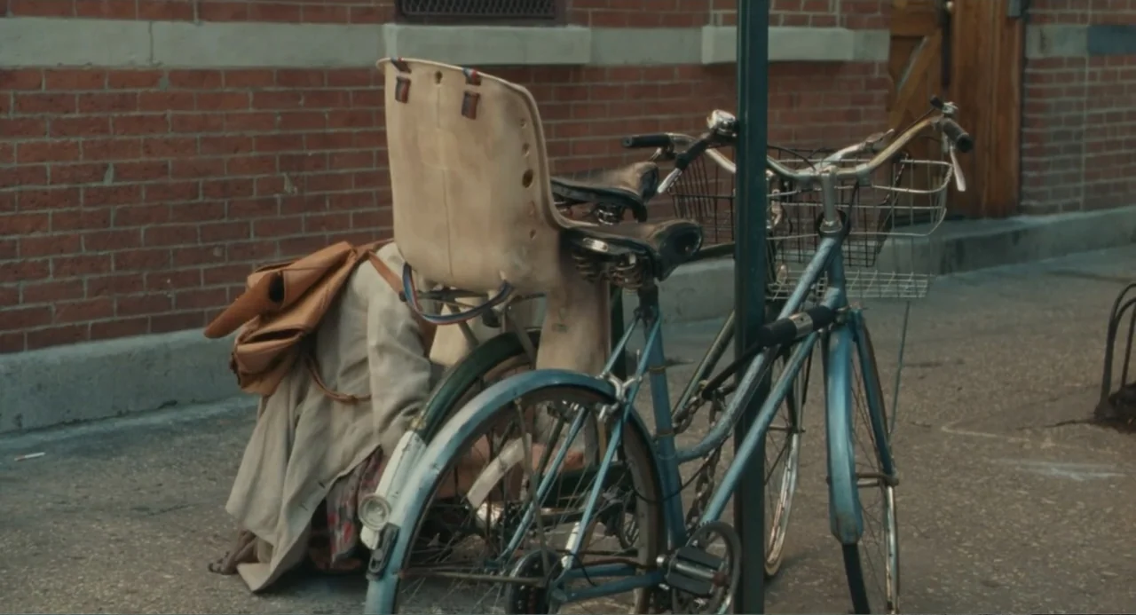 Motherhood (2020) movie still: medium — A sidewalk sits adjacent to a weathered red brick wall. A person in a beige coat crouches beside a light…