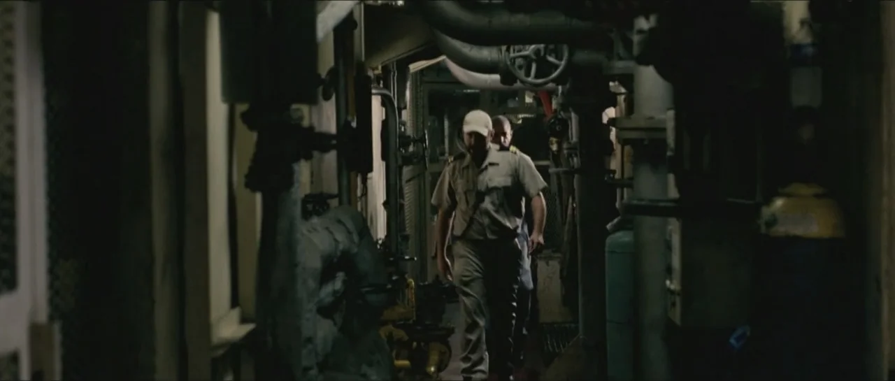Contraband (2012) movie still: fullbody — A narrow corridor inside a metal vessel is filled with pipes and industrial hardware. A man in a uniform…