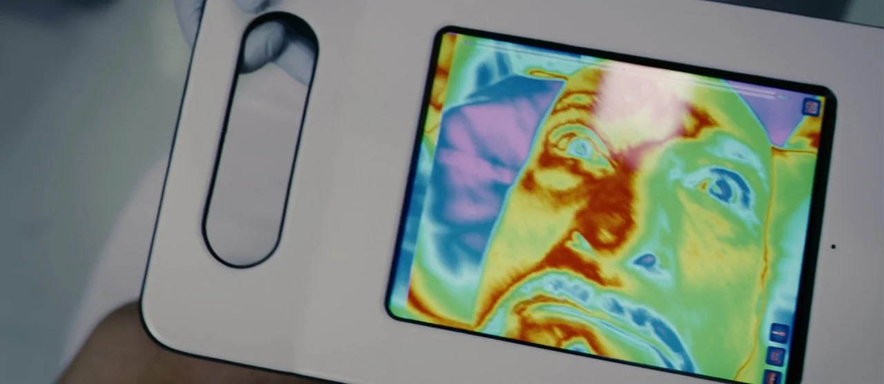Self/less (2015) movie still: closeup — A white handheld diagnostic device displays a thermal imaging scan of a human face. A gloved hand holds the…