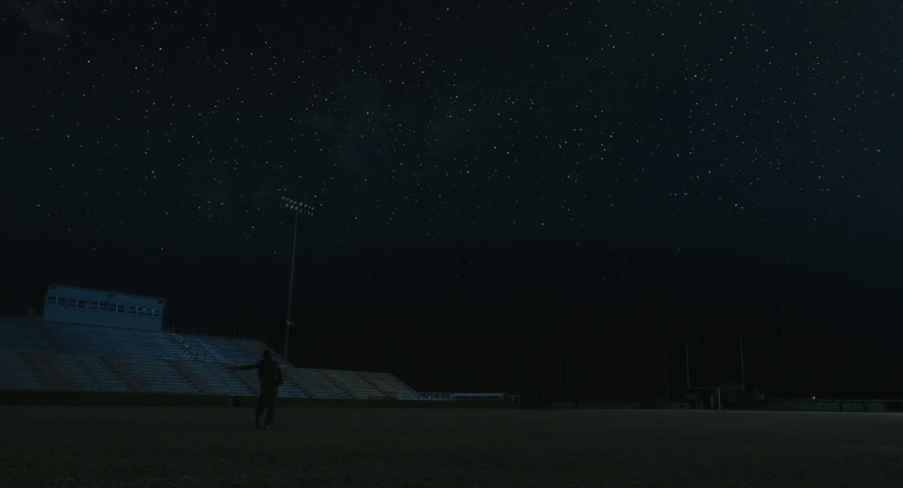 The Life of Chuck (2024) movie still: wide — A vast, empty high school football stadium under a clear star-filled night sky. A lone figure stands on…