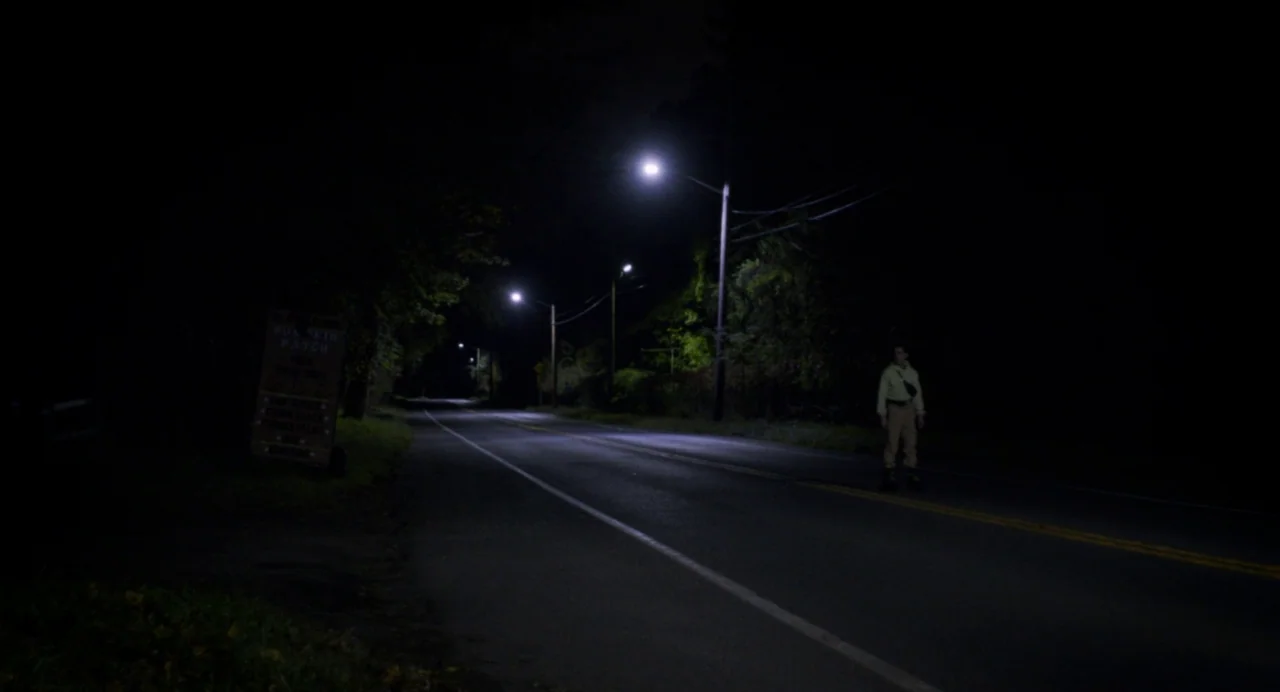 Happer’s Comet (2022) movie still: wide — A desolate road at night features glowing streetlamps and a wooden sign board. A solitary figure stands on…