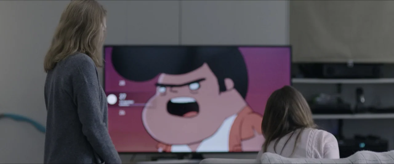 The Lie (2018) movie still: medium — A living room interior contains two people seated before a large flat-screen television. The woman and child…