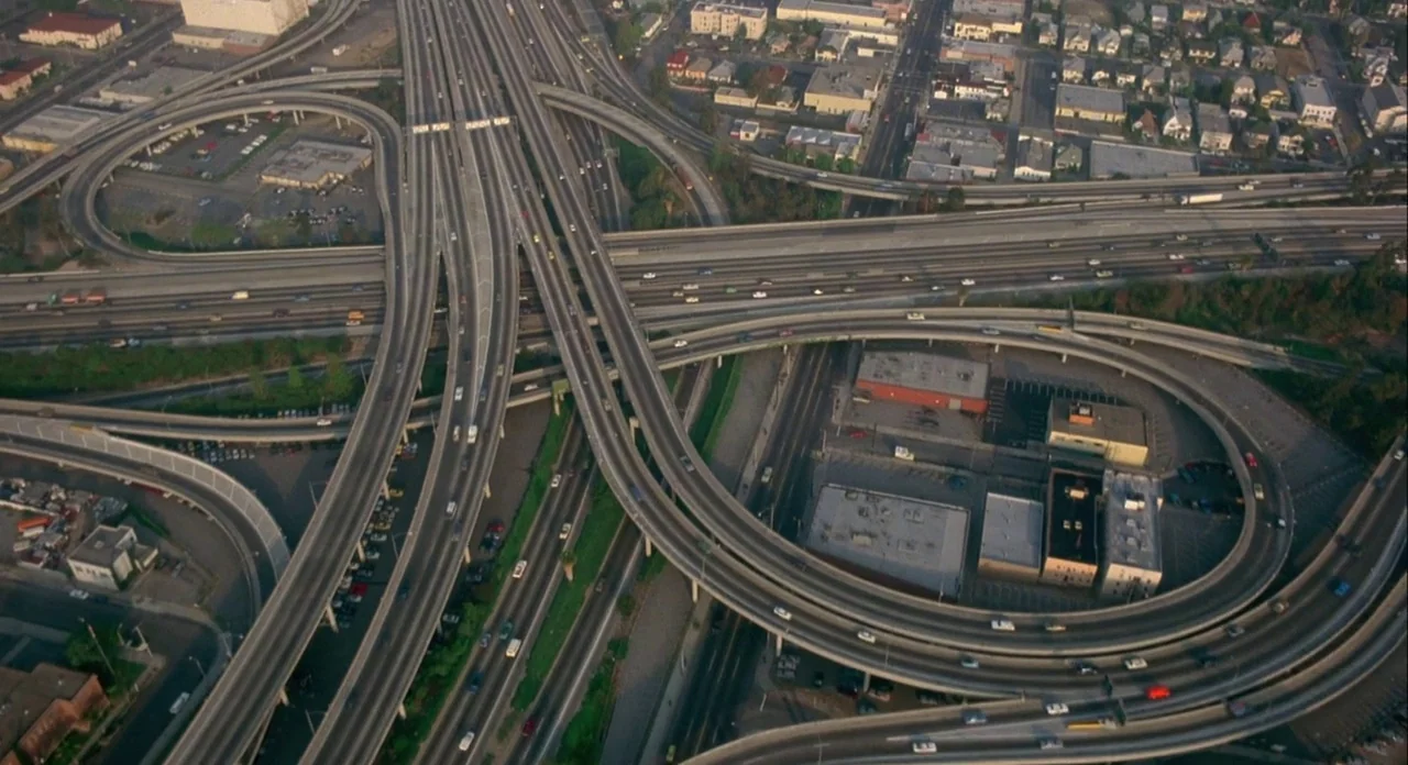 Koyaanasquatsi (1982) movie still: establishing — An aerial view captures a sprawling concrete highway interchange with multiple curving overpasses.…