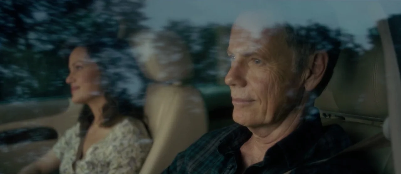 Gerald’s Game (2017) movie still: medium — Inside a moving car with reflections of passing trees visible on the window glass. An older man in a…