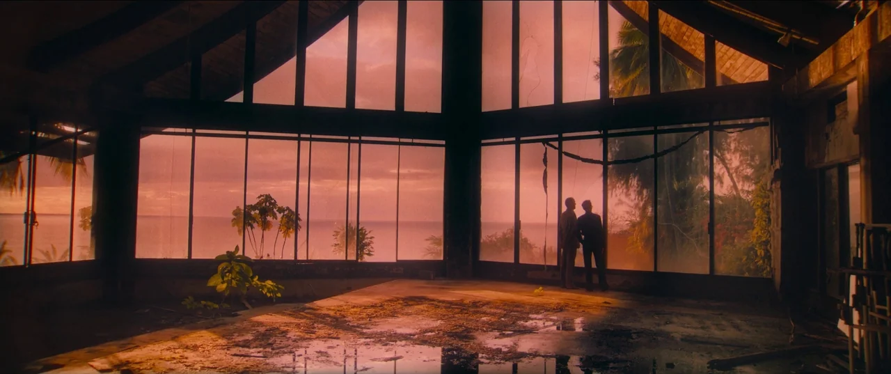 Pacifiction (2022) movie still: wide — An abandoned open-plan structure overlooks a tropical ocean at sunset through massive glass windows. Two men…