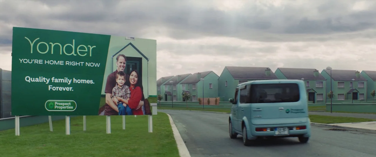 Vivarium (2019) movie still: wide — A suburban street lined with uniform green houses under a cloudy gray sky. A large billboard featuring a family…