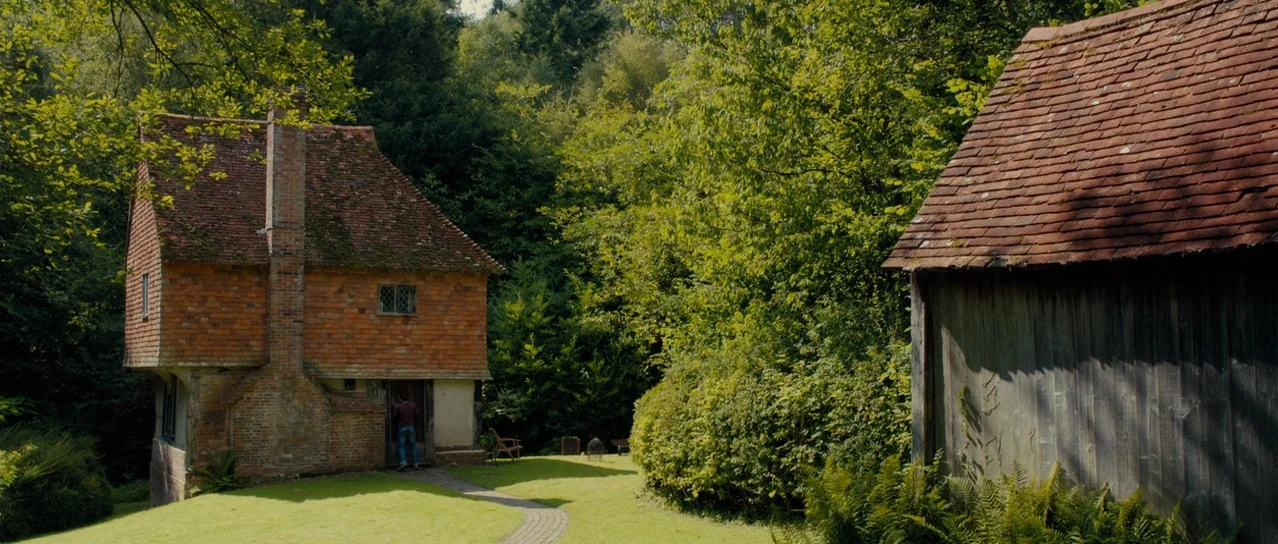 Road Games (2015) movie still: wide — A traditional brick house with a tiled roof sits in a wooded clearing. A person stands at the doorway of the…