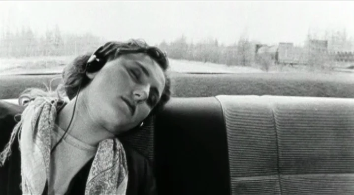 Roadkill (1989) movie still: medium — The interior of a car backseat with fabric upholstery is visible. A woman with headphones sleeps against the…