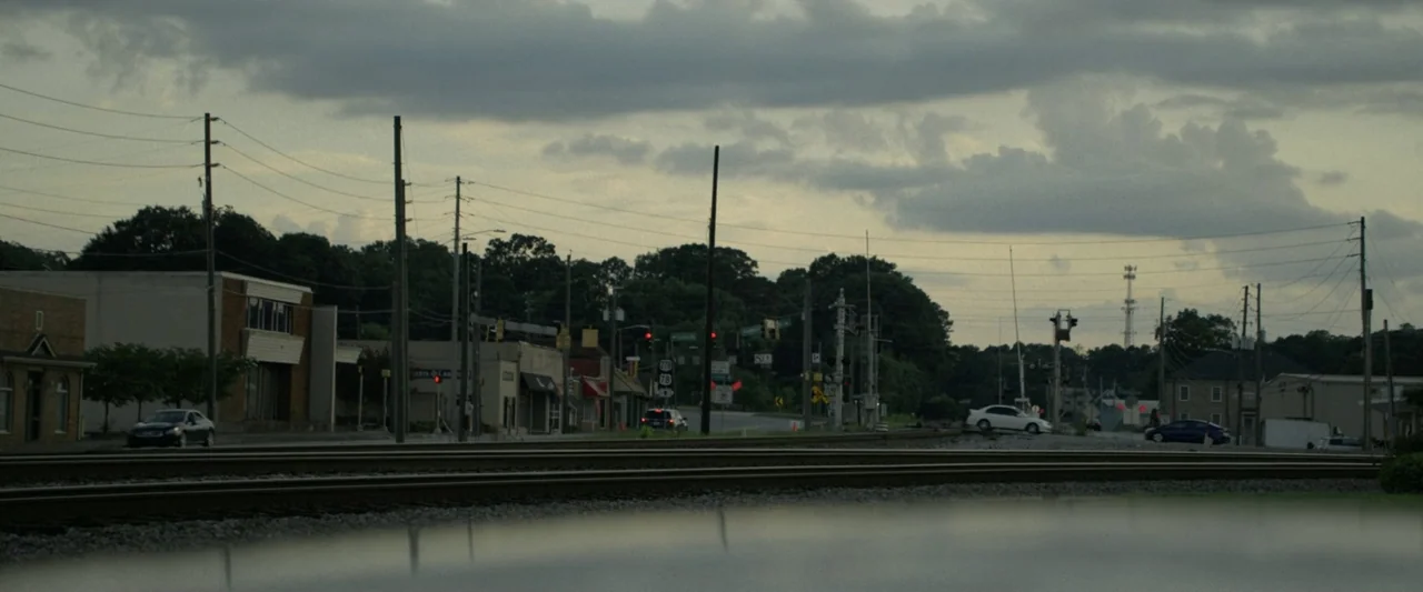 One Fast Move (2024) movie still: wide — A quiet road intersection features railroad tracks running horizontally across the frame foreground.…