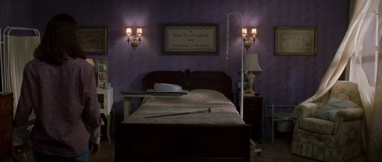 The Haunting (1999) movie still: medium — A bedroom with dark purple patterned wallpaper and a central wooden bed frame. A woman stands in the…