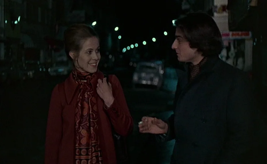 Love on the Run (1979) movie still: medium — A dark, dimly lit city street at night with distant bokeh lights. A young woman and man are positioned…