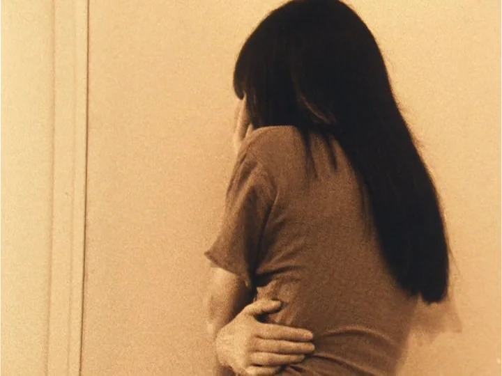 The Woman with Two Heads (1977) movie still: medium — A person stands against a plain light-colored wall in a simple interior space. The subject has…