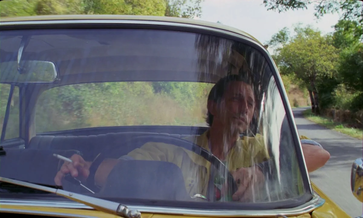 Betty Blue (1986) movie still: medium — A man drives a vintage yellow car along a tree-lined rural road. He holds a cigarette in his right hand…