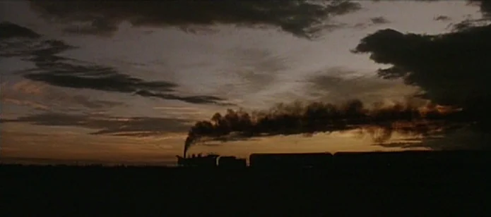 The Neon Bible (1995) movie still: wide — A steam locomotive travels across a wide, flat landscape under a dark, dusk sky. Thick, black exhaust…