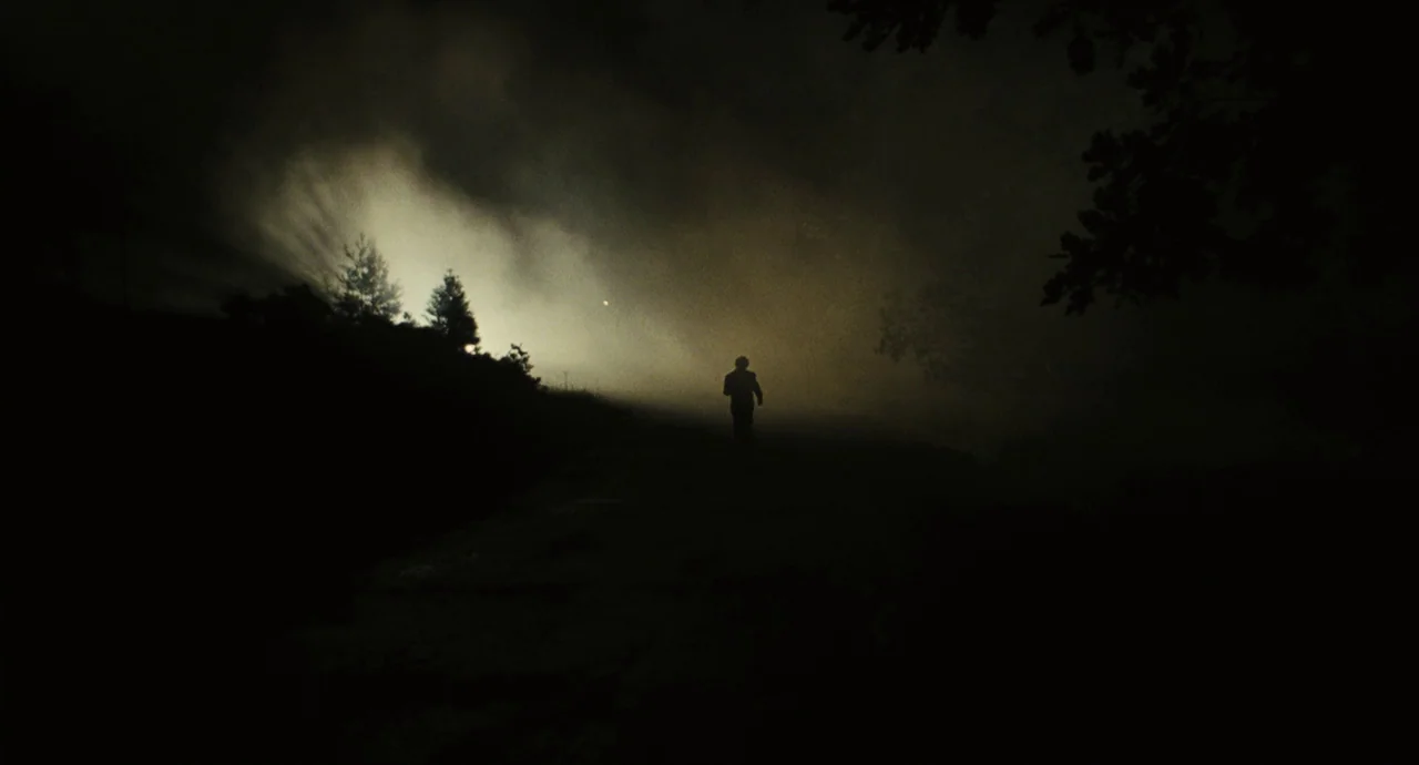 The Double (2013) movie still: wide — A wide, dark field is filled with heavy fog and silhouetted trees at night. A lone figure walks away from the…