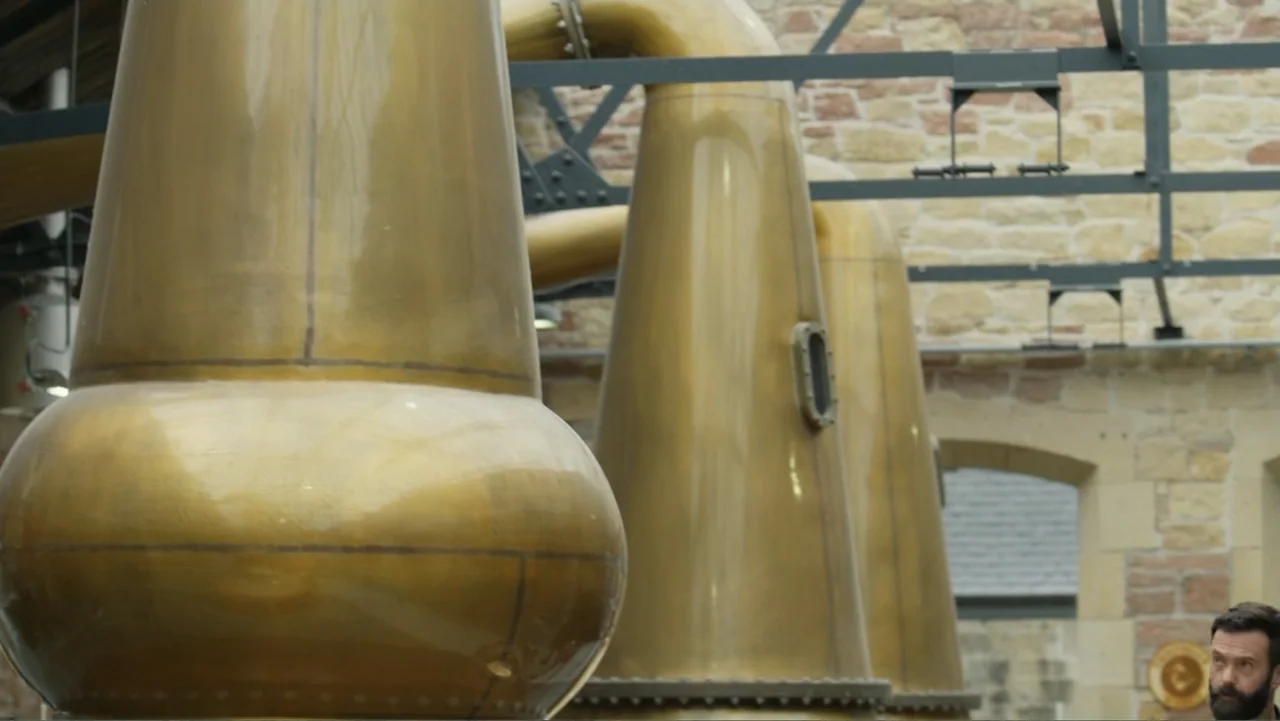 A Scottish Love Scheme (2024) movie still: medium — Large polished copper distillation stills dominate the foreground of a stone-walled facility. A…
