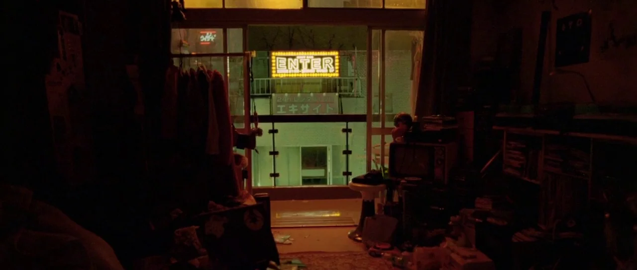 Enter The Void (2009) movie still: wide — A dark, cluttered apartment interior looks out onto an illuminated urban exterior at night. Neon signs…