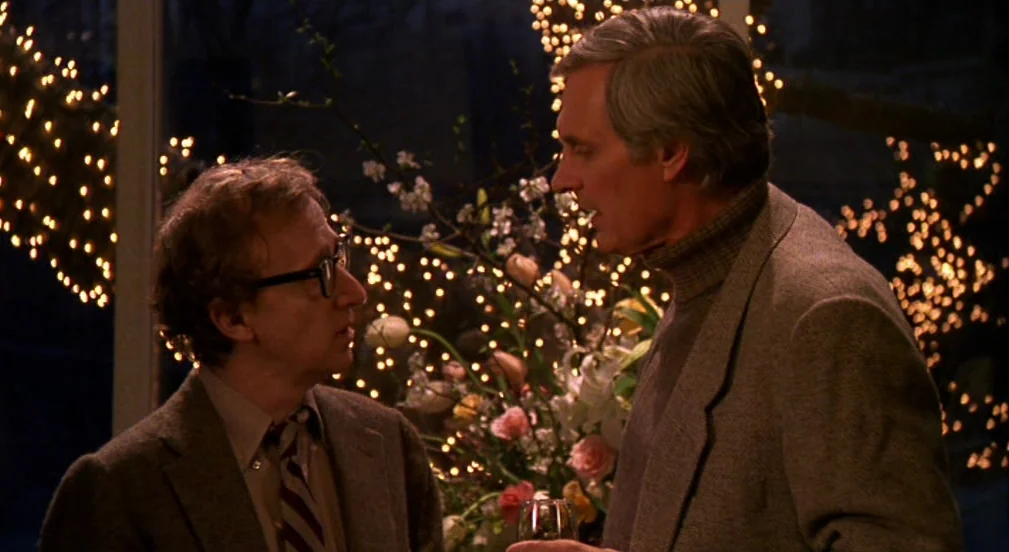 Crimes and Misdemeanors (1989) movie still: medium — An interior space features festive string lights blurred in the background behind two men. The…