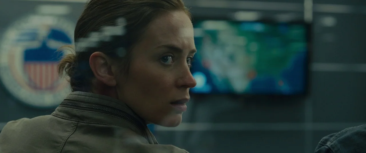 Sicario (2015) movie still: closeup — A dimly lit room features a government emblem on the wall behind a monitor. A woman turns her head to look…