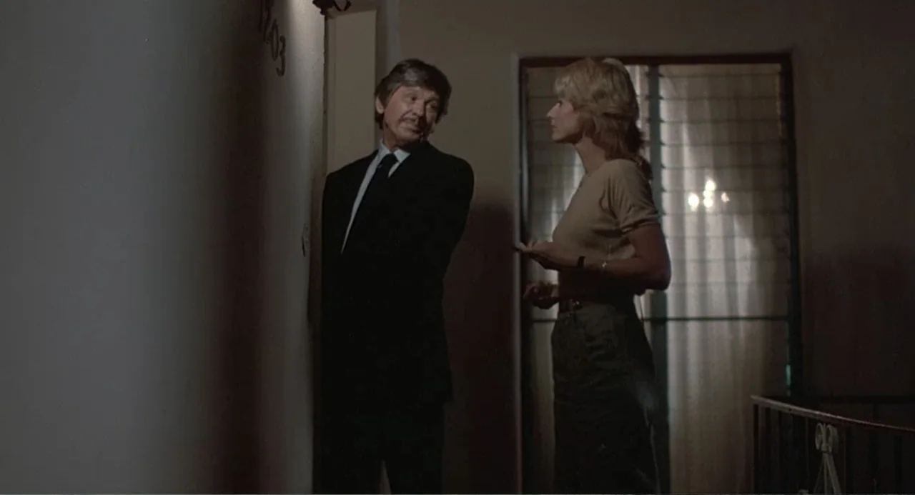 Death Wish II (1982) movie still: medium — A dimly lit hotel corridor features a plain wall with a visible room number. A man in a suit stands by…